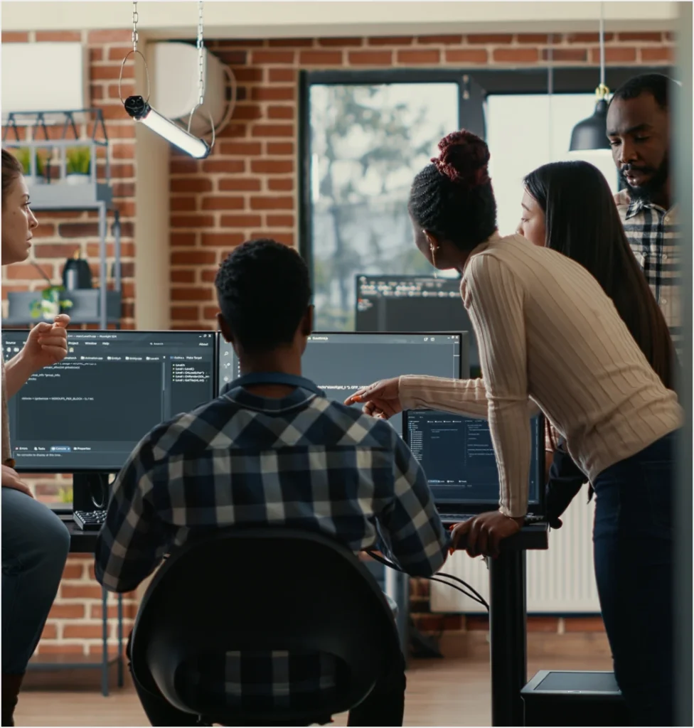 Diverse team of software developers collaborating around computer monitors displaying code in a modern industrial office space.