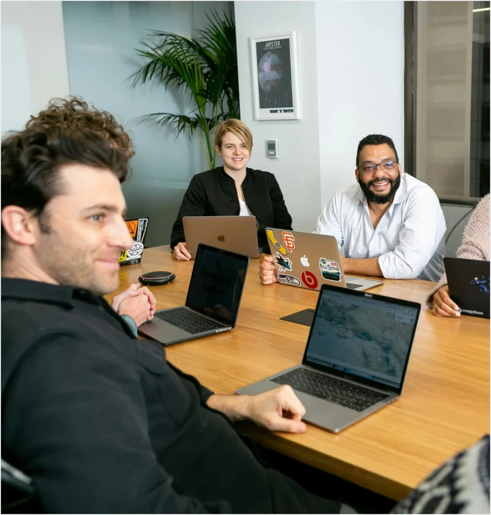 Four professionals collaborating around a wooden conference table, each with a laptop open. The diverse team includes three men and one woman, all engaged in a productive work session in a modern office setting with natural lighting, plants, and contemporary decor.